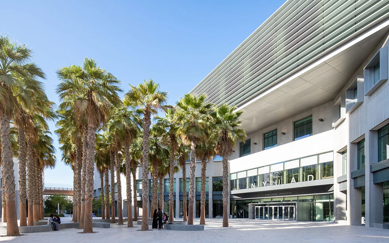The Campus Center at NYU Abu Dhabi, its louvred upper floor projecting over a glass entrance, with an avenue of palms and students sitting on low benches in front of it.