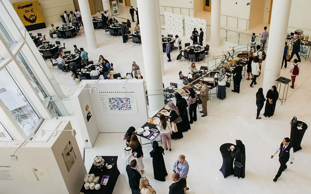 A conference reception in a double-height atrium on campus: round tables laid for lunch, a buffet line, and people standing among display boards.