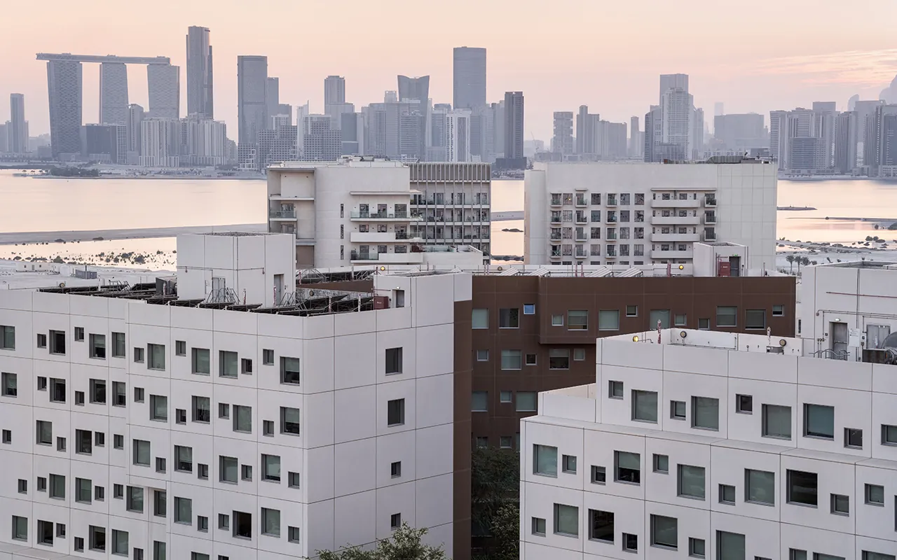 Campus residential buildings at dusk, with the towers of central Abu Dhabi across the water behind them.